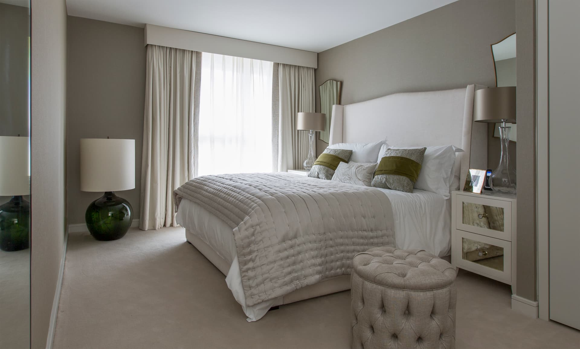 Pale principal bedroom with a tall upholstered headboard and tufted stool.