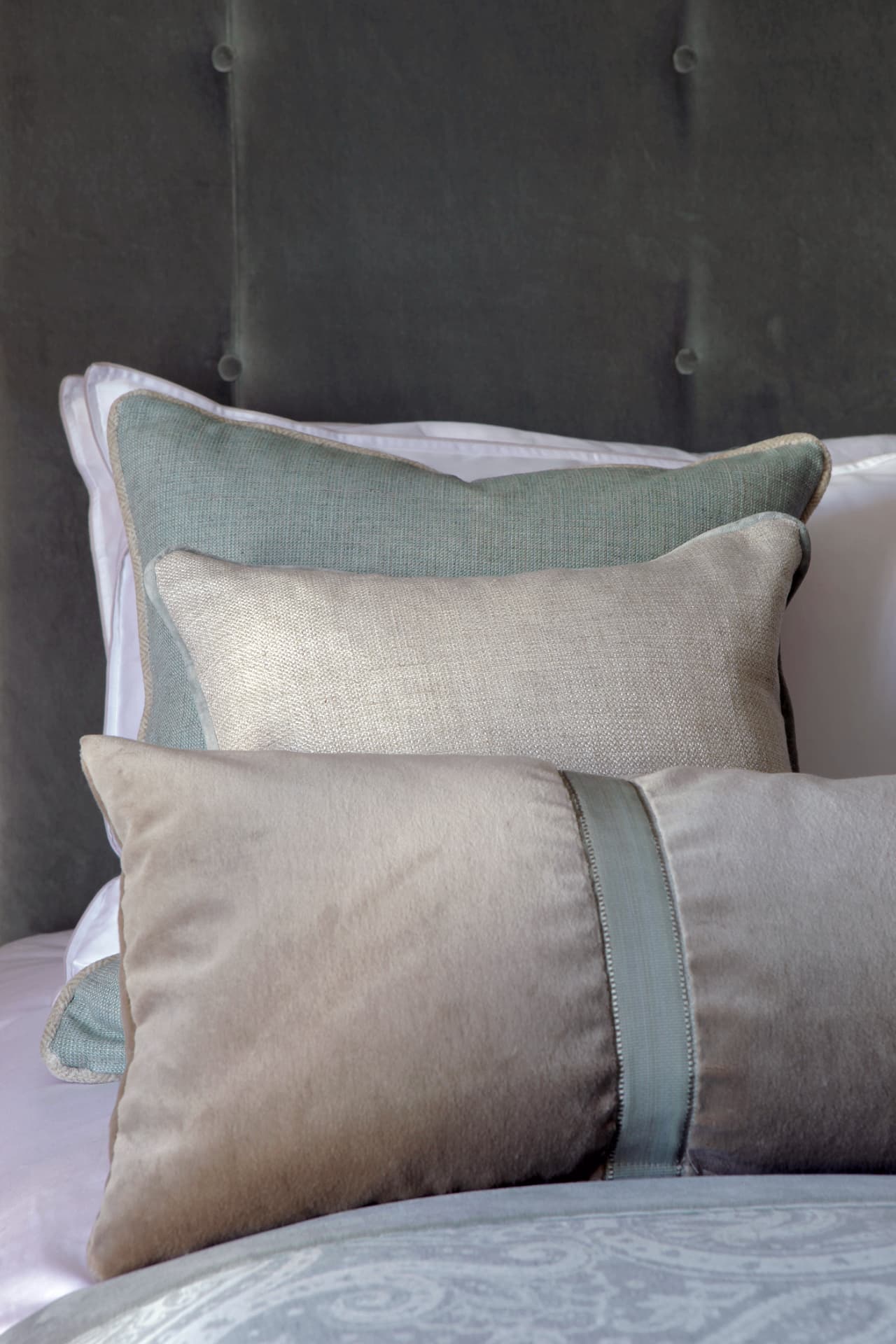Layered cushions in soft grey-green, stone and blush linens against an upholstered headboard.