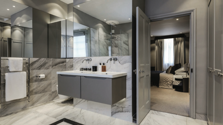 A grey-tiled bathroom with marble surround, twin vanity unit and a view through an open door into a bedroom.